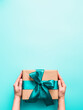Female hands hold big gift box on turquoise blue background, copy space. Caucasian girl hands holding gift box in craft wrapping paper with green satin ribbon. Christmas, New Year or Birthday present