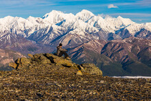 A Hiker Resting On Rocks Observes McGinnis Peak, Mount Moffit, And Mount Hayes Towering In The Distance; Alaska, United States Of America