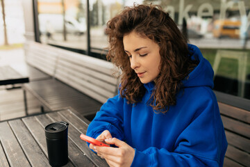 Close up outside photo of stylish lovely woman with wavy hair wearing blue oversize shirt looking in smartphone and chatting with friends. Girl resting outdoor in city cafe. 