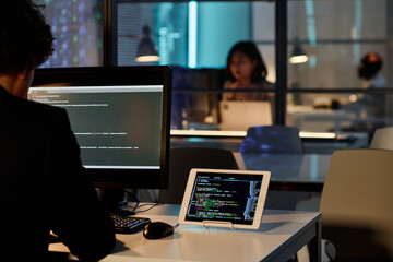 Rear view of developer sitting at table in front of computer monitor and rewrite codes from tablet pc on computer