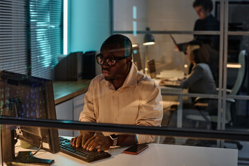 African American developer typing on keyboard and working on computer with software till late at night with colleagues in background