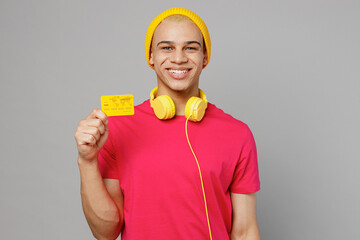 Wall Mural - Young smiling happy fun cheerful man of African American ethnicity 20s he wearing pink t-shirt yellow hat headphones hold in hand mock up of credit bank card isolated on plain grey background studio.