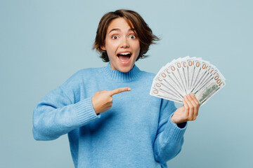 Wall Mural - Young surprised woman wear knitted sweater look camera hold point finger on fan of cash money in dollar banknotes isolated on plain pastel light blue cyan background studio. People lifestyle concept.