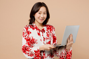 Wall Mural - Young happy smart IT woman of Asian ethnicity wear red shirt look camera hold use work on laptop pc computer isolated on plain pastel light beige background studio portrait. People lifestyle concept.