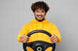 Young cool happy cheerful Indian man 20s he wearing casual yellow hoody hold steering wheel driving car pretend to drive isolated on plain grey background studio portrait. People lifestyle portrait.