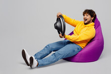 Full Body Excited Fun Young Indian Man 20s He Wearing Casual Yellow Hoody Sit In Bag Chair Hold Steering Wheel Driving Car Isolated On Plain Grey Background Studio Portrait. People Lifestyle Portrait.