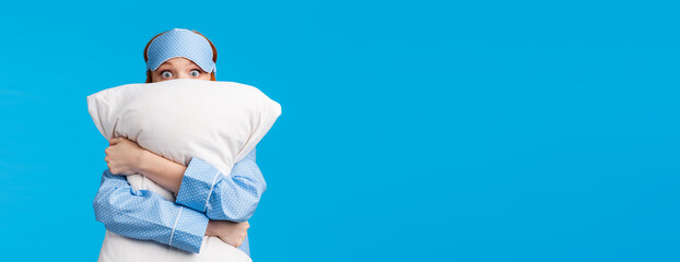 Wall Mural - Silly scared young timid glamour redhead girl feeling scared waking up night strange frightening noise, hugging pillow, peeking with horrified eyes, wearing sleepwear and sleep mask, blue background