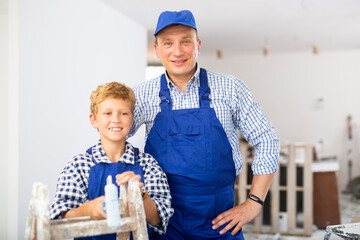 Wall Mural - Portrait of positive father and son wearing blue overalls, standing in construction site in apartment.