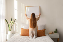 Young Woman Hanging Blank Frame On Light Wall In Bedroom, Back View