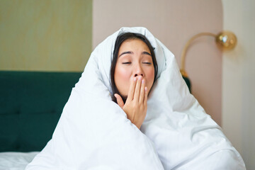 Close up of funny sleepy girl, asian woman yawns after waking up early morning, covers herself with blanket duvet, holds hand near opened mouth