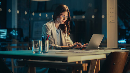 Office at Night: Focused Female Manager Working Long Hours Alone in Creative Agency, Implementing Business Strategy for Client. Creative Specialist Writing Corporate Project Plans on Laptop Computer.