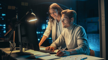 Wall Mural - Software Developer Working on Desktop Computer in the Evening in Office. Young Female Project Manager Comes Over to the Desk to Discuss Updates to the New Online Application.