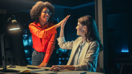 Wall Mural - Art Director and Graphic Designer Discussing Marketing Campaign for Business Partner. Two Young Successful Women High Five Each Other. Small Creative Team Working in the Evening in Office.