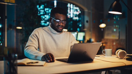 Poster - Stylish Black Specialist Sitting at a Desk in Creative Agency in the Evening. Young Handsome African Man Working on a Laptop Computer in Marketing Company. High Angle Footage.