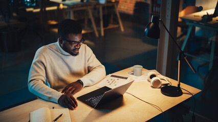 Poster - Handsome Manager Sitting at a Desk in Creative Office. Young Stylish Black Man in White Jacket Using Laptop Computer in Marketing Agency in the Evening. High Angle Footage.