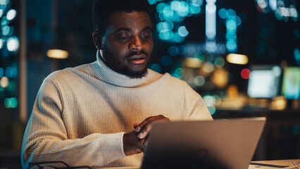 Poster - Handsome Black Project Manager Making a Video Call on Laptop Computer in an Evening Office Environment. African Specialist Talking to Colleagues Over a Live Camera, Working Remotely on a Project.