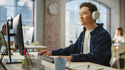 Fototapete - Portrait of Asian Creative Focused Man Smiles While Using Computer and Headphones in Bright Modern Office. Manager Planning a Project While Listening to Podcast.