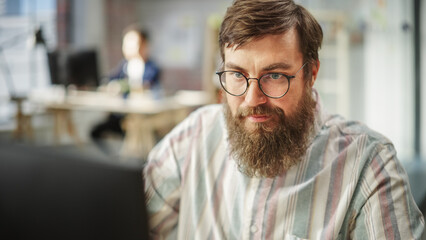 Poster - Portrait of Young Caucasian Bearded Man with Glasses Focused and Working on Computer in a Bright Spacious Office. Male Administrator Smiling after Successfully Finishing a Task.