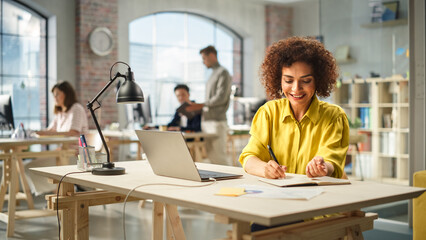 Fototapete - Portrait of Biracial Active Young Woman Working on Laptop in Busy Office. Biracial Female Manager smiling while Taking Notes one a Notebook From Her Laptop . People Using Computers in the Background.