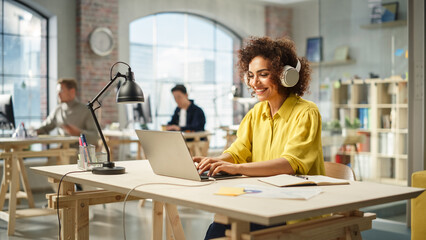 Fototapete - Portrait of Biracial Young Woman Smiling and Using Laptop in Bright Contemporary Office. Client Advisor Answering Customer Support Through Headphones and Typing. Team Working in the Background