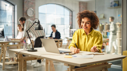 Poster - Portrait of Young Biracial Woman Taking Notes at Her Desk While Looking for References on the Laptop. Busy Employees Are Collaborating in the Background.