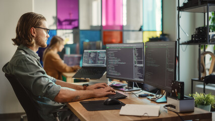 Wall Mural - Professional Male Developer Writing Code On Desktop Computer With Two Displays and a Laptop in Creative Office. Caucasian Man Programming Artificial Intelligence Software For Big Tech Company.