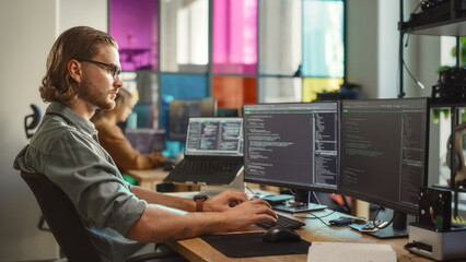 Wall Mural - Young Caucasian Man Coding on Personal Computer and Laptop Set Up In Stylish Office. Professional Programmer Developing Innovative AI Software in Technological Start-Up, Using Multiple Displays.