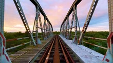 POV Of Walking On Iron Bridge At Sunset. Abandoned Old Railway Bridge. Rusty Metal Bridge With The Sky In The Colors Of The Sunset In The Background. 