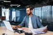 © Liubomir - A serious young man accountant, financier, analyst, auditor sits in the office at the table. He holds documents and a pen in his hands, checks accounts, finances, types on a laptop.