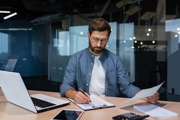 A serious young man accountant, financier, analyst, auditor sits in the office at the table. Works with documents. writes and calculates the budget, finances.