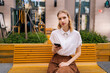 Portrait of confident charming young woman sitting on bench and looking at camera holding mobile phone in hand on background of building. Serious female using mobile phone outdoors in summer.