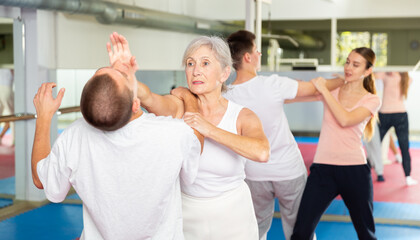 Wall Mural - Senior European woman learning chin strike move on man during self-defense training. Othe woman training with young man in background.