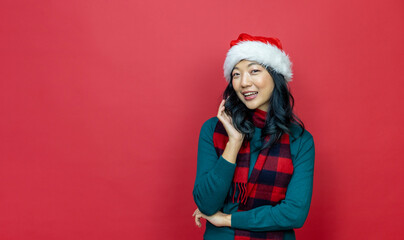 Wall Mural - Pretty smiling asian woman in warm christmas sweater and santa hat on red background for season celebration concept