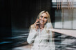 © Iona - Beautiful blonde businesswoman in white suite talking by phone, broad smiling, looking up through the window on sunny day, excited by good news. Happy Scandinavian woman at work, loving her job.