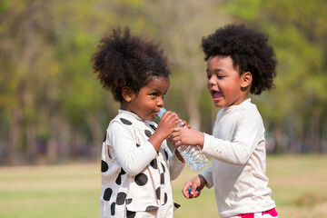 Two little children drinking water while playing together outdoor in the park. Child girl and boy having fun outdoors. Black kid people enjoying outside