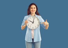 Portrait Of Beautiful Happy Young Brunette Holding Round Wall Clock In One Hand And Pointing Her Finger At Them With The Other Hand. Studio Photo On Isolated Blue Background. Time Concept.