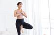 © artitwpd - Beautiful happy young Asian woman standing in Vrksasana Posture, doing yoga exercise on purple mat with water bottle, working out in bedroom, indoor workout, Tree Pose.