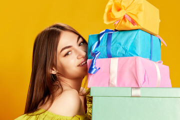 Portrait of young beautiful girl in cute dress posing with many present boxes over yellow background. Surprise, gifts