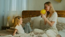 Caucasian Adult Mother With Daughter Sitting Playing On Bed In Bedroom Mum Blowing Yellow Balloon Blow Air In Inflatable Ball Prepare Festive Birthday Event Little Girl Child Kid Having Fun With Mom