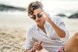 Portrait of handsome young italian man relaxing at the beach, wearing white shirt and fashionable sunglasses.