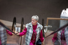 Fashionable Stylish Aged Woman With Gray Hair Standing On Escalator, Wearing Bright Look Of Plaid Vest Jacket, Burgundy Dress