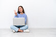 © luismolinero - Young caucasian woman with laptop sitting on the floor isolated on white background smiling and showing victory sign