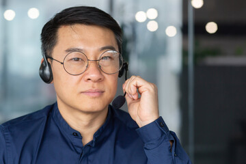 Close up photo. Portrait of a serious Asian man in a headset. He looks into the camera, holds the microphone in his hand. Businessman, business trainer, saleman, customer support hotline.