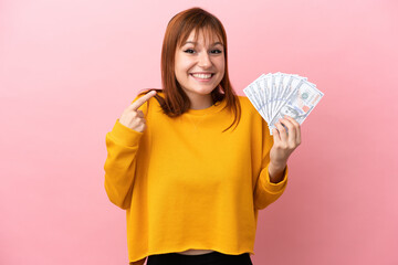 Wall Mural - Redhead girl taking a lot of money isolated on pink background giving a thumbs up gesture