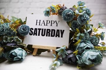 Poster - Happy Saturday text on wooden easel standing on white brick wall and wooden background