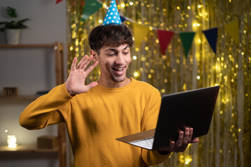 Virtual celebration. Portrait of happy young arabic man wearing party hat holding laptop and waving to webcam at home interior, making online video call on computer, greeting with birthday