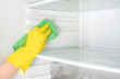 Hand cleaning refrigerator. Person washing refrigerator with rag. Housekeeper wipes shelves of clean refrigerator. Hand in yellow rubber protective glove and green sponge washes. copy space