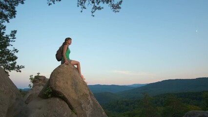 Wall Mural - Young woman hiker sitting alone on rocky mountain enjoying view of evening nature on wilderness trail. Active way of life concept