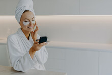 Beautiful Happy Young Lady Applies Collagen Patches Under Eyes Looks At Herself In Mirror Does Rejuvenation Procedure Wears Bath Towel And Dressing Gown After Taking Shower Has Skin Care Routine