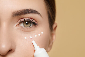 Sticker - Young woman applying cream under eyes on beige background, closeup. Space for text